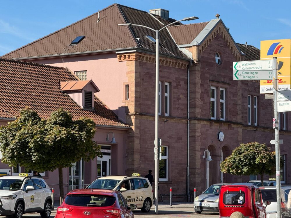 Pink brick building with red-tiled roof and street signs, with taxis parked along the curb on a sunny day in a European town center.