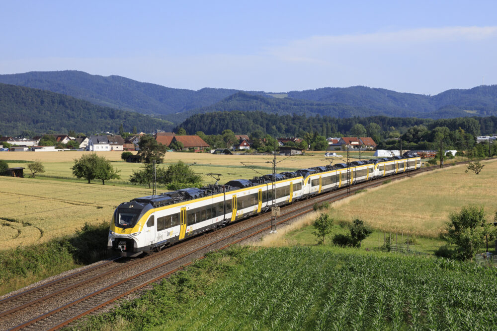 Modern yellow-and-white passenger train travels along a double-track through a rural landscape with a village, fields, and forested hills in the distance under a clear blue sky.