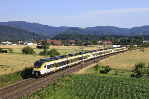 Modern yellow-and-white passenger train travels along a double-track through a rural landscape with a village, fields, and forested hills in the distance under a clear blue sky.