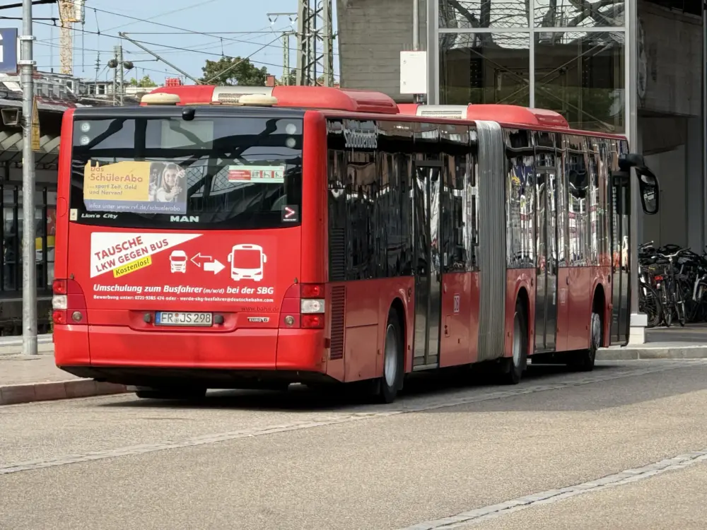Red public bus parked at a curb with gold and white ads on the front window and a city street backdrop behind it.