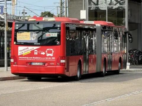 Red public bus parked at a curb with gold and white ads on the front window and a city street backdrop behind it.